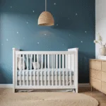 Een fotorealistische weergave van een serene Scandinavische kinderkamer met zachte, natuurlijke zonneschijn die door hoge ramen stroomt. De kamer bevat een witte houten wieg met lichtblauw en grijs beddengoed, een natuurlijke eiken ladekast, een jute vloerkleed en een opvallende petrolblauwe accentmuur met delicate sterrenpatronen. Een rieten hanglamp hangt boven, terwijl de strakke witte muren een minimalistisch Noordisch ontwerp accentueren. De foto is genomen op ooghoogte tijdens het gouden uur, met een subtiele scherptediepte en prominente textuurdetails in hoge resolutie.