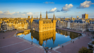Aerial view of The Hague's Binnenhof complex reflecting in Hofvijver pond, showcasing Gothic architecture, modern government buildings, and tourists in the courtyard under golden afternoon light with Dutch flags flying against a partly cloudy blue sky.
