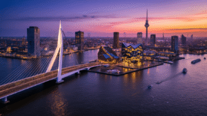 Aerial view of Rotterdam at dusk featuring the illuminated Erasmus Bridge, Cube Houses, and Markthal, with the Euromast tower in the background and boats on the Maas River, showcasing a blend of historic and modern architecture against a vibrant purple-orange sky.