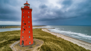 De Texelse Vuurtoren Die 72 Schepen Had Kunnen Redden (En Waarom Je Hem Móét Beklimmen) Dramatic wide-angle view of Eierland lighthouse on Texel, Netherlands, showcasing its iconic red brick structure against stormy skies, highlighting bullet holes from WWII and the surrounding landscape of Wadden Sea and North Sea, emphasizing its historical significance.